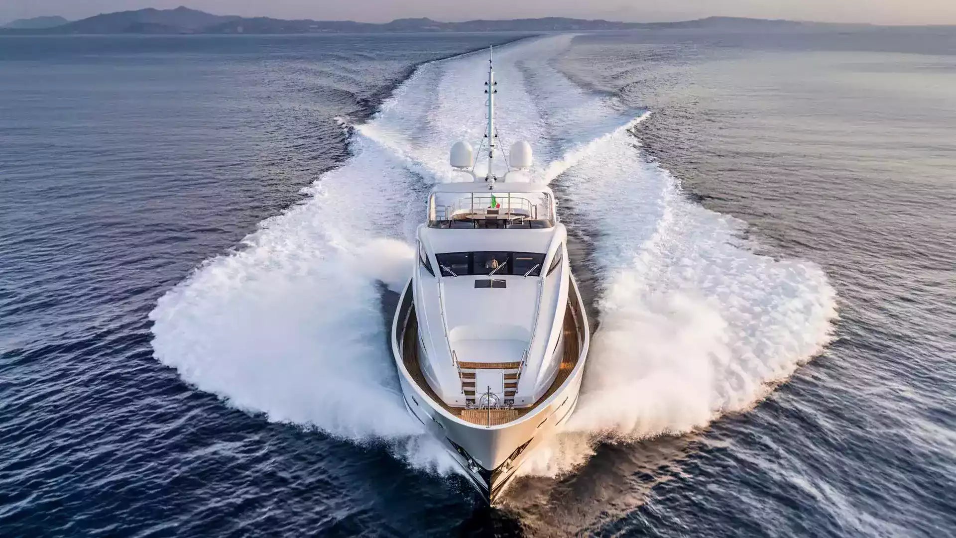 A luxury motor yacht speeds across deep blue ocean water, creating a large white wake behind it under a clear sky.