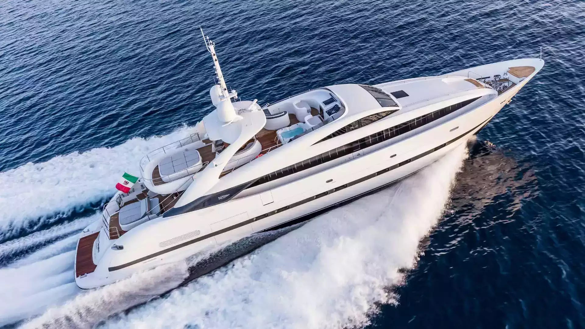 A luxury white motor yacht with brown wood accents sailing at an angle on deep blue water, creating a white wake behind it.