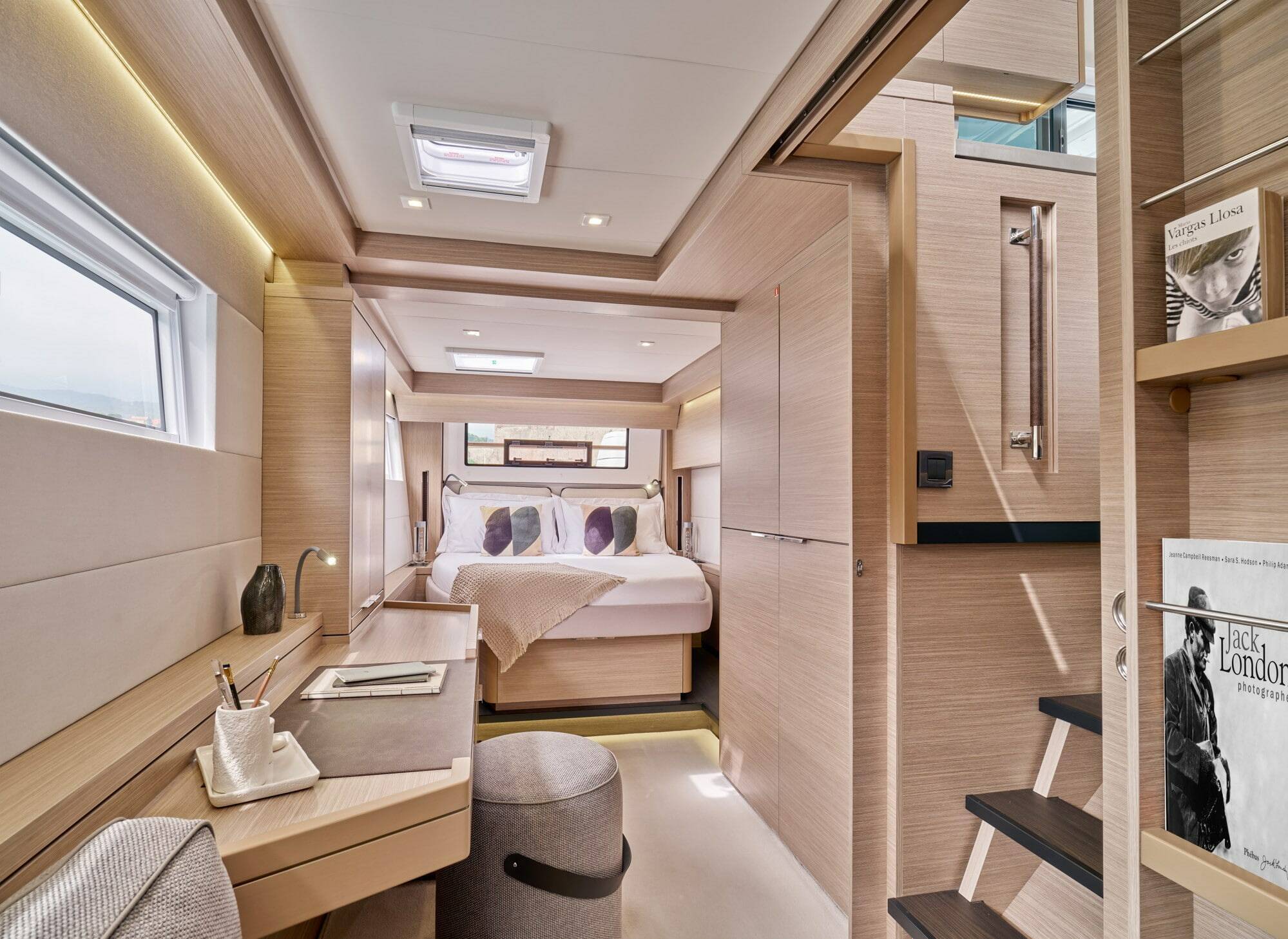 A luxury yacht or motorhome interior featuring a spacious bedroom with a neatly made bed, a built-in desk with a vanity mirror, a gray cylindrical stool, and a staircase on the right side.