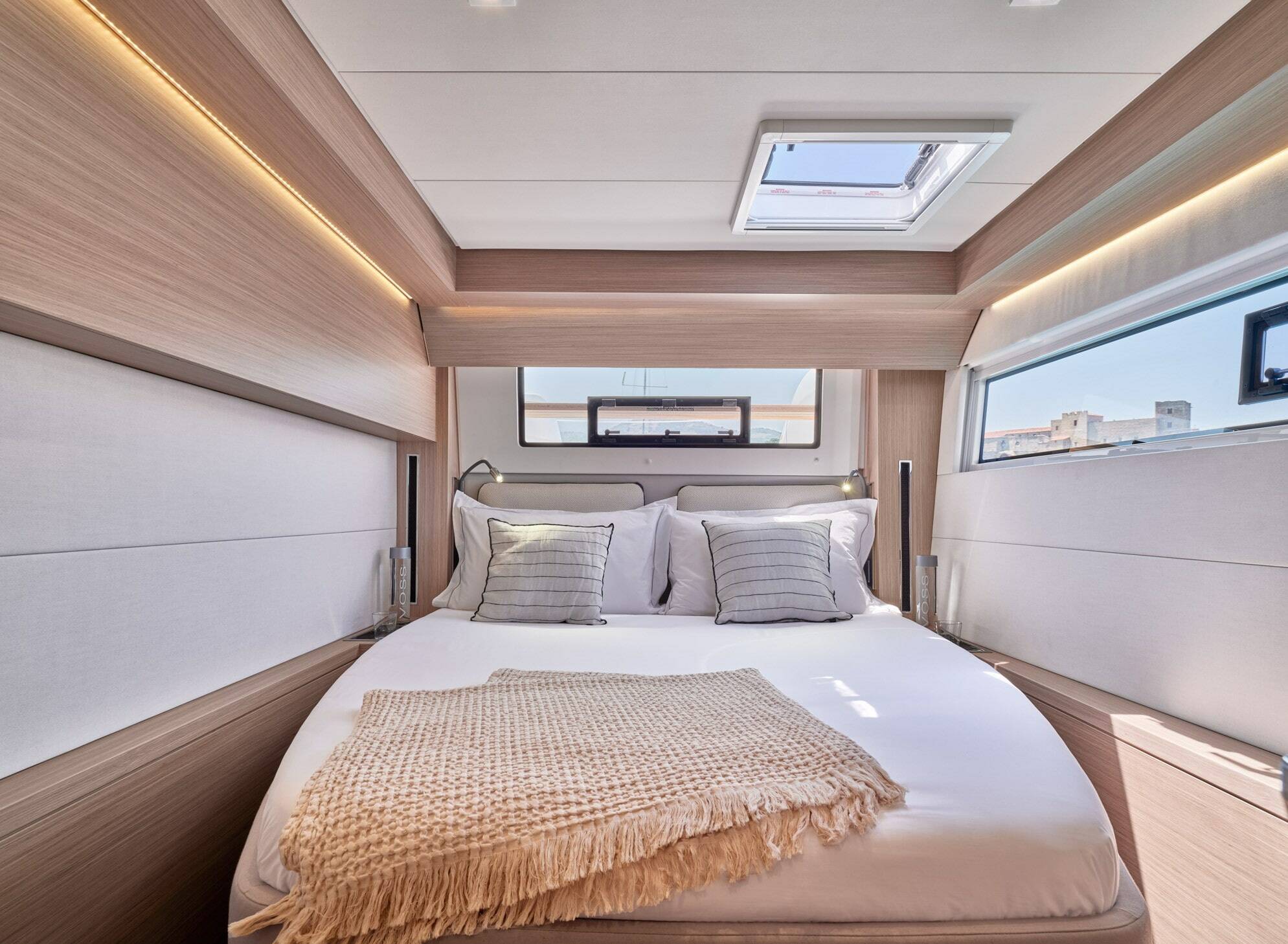 A modern yacht or boat cabin bedroom featuring a neatly made bed with white linens, striped pillows, and a beige woven throw blanket. The room has light wood paneling, built-in LED lighting, and large windows looking out onto water.