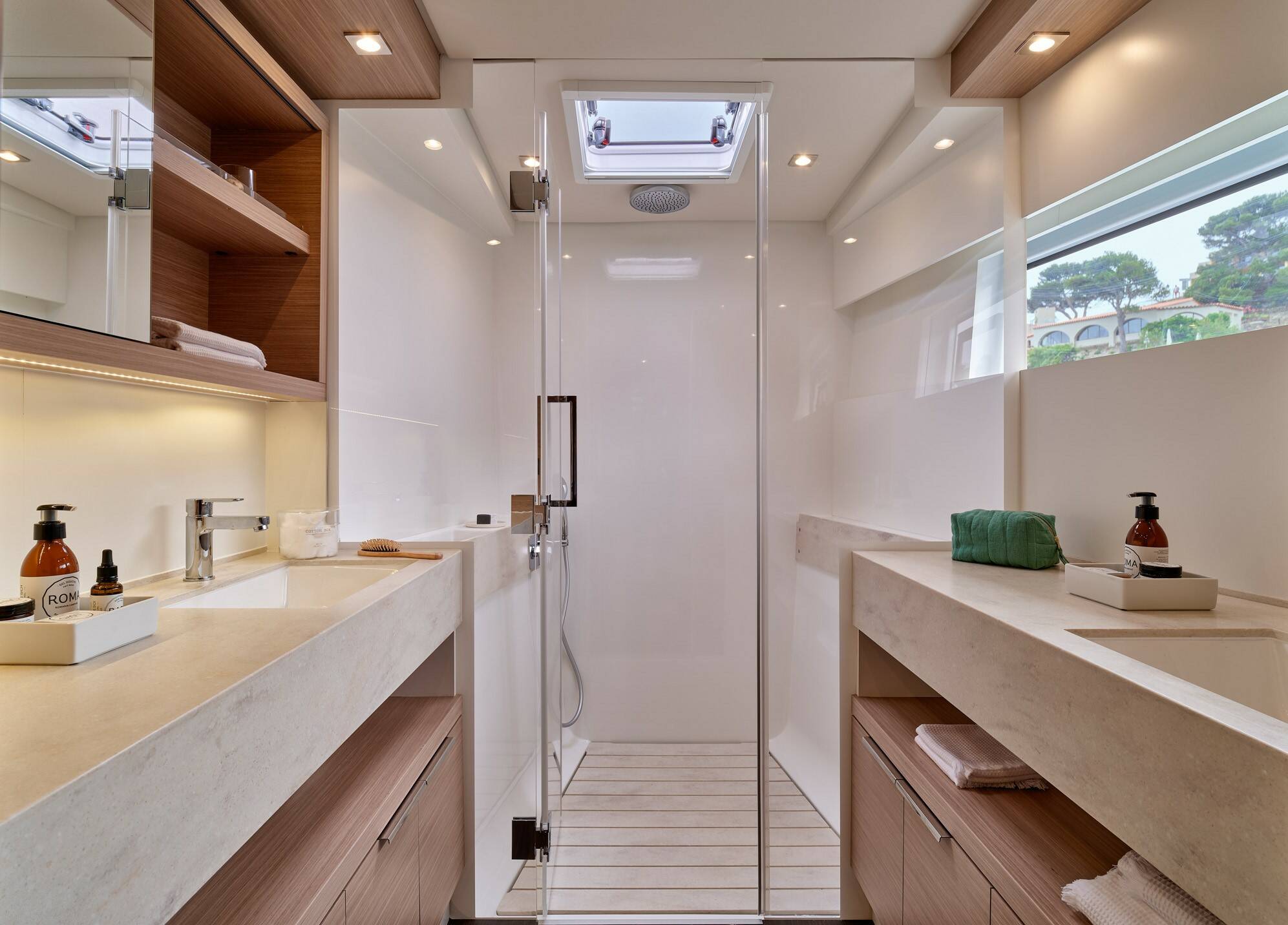 A modern yacht bathroom featuring a walk-in glass shower enclosure, double vanity counters with light stone tops, wooden cabinetry, and a bright, airy interior.