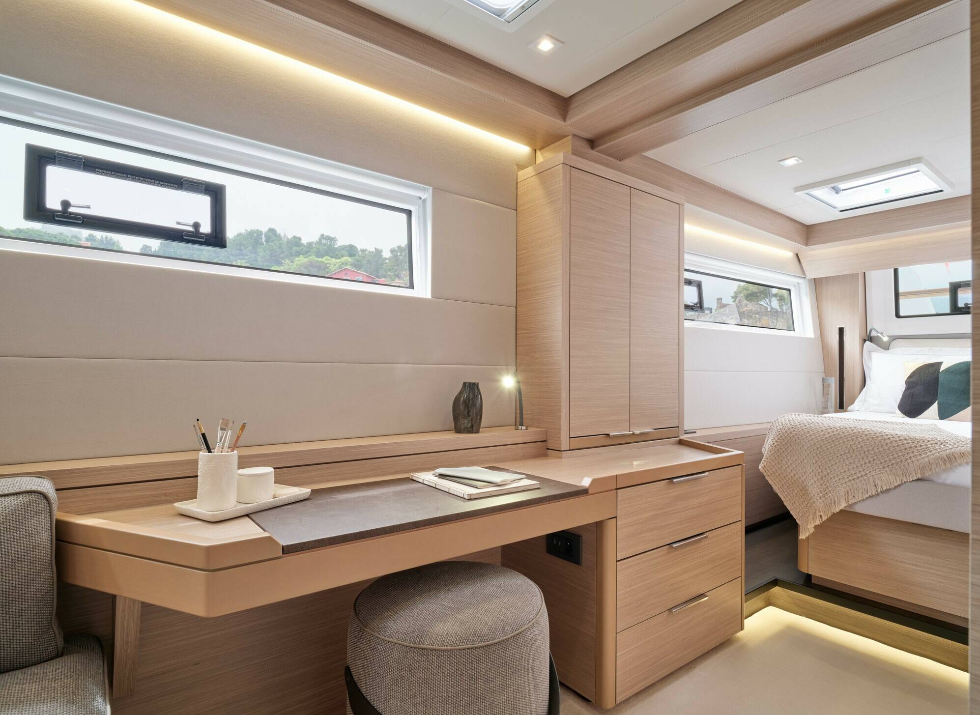A modern yacht or cabin interior featuring a light wood vanity desk with drawers, a gray upholstered stool, built-in cabinets, and a bed area with a cream quilt in the background.