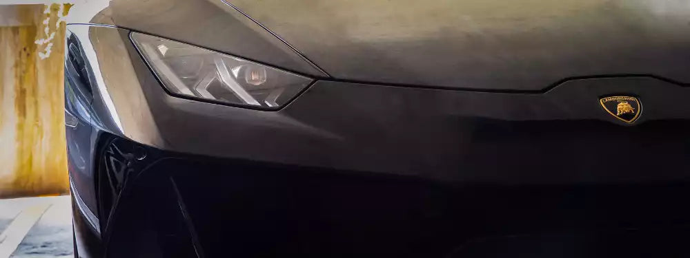 Close-up of the front end of a dark metallic Lamborghini sports car, showing the Lamborghini bull emblem on the hood and part of the headlight.
