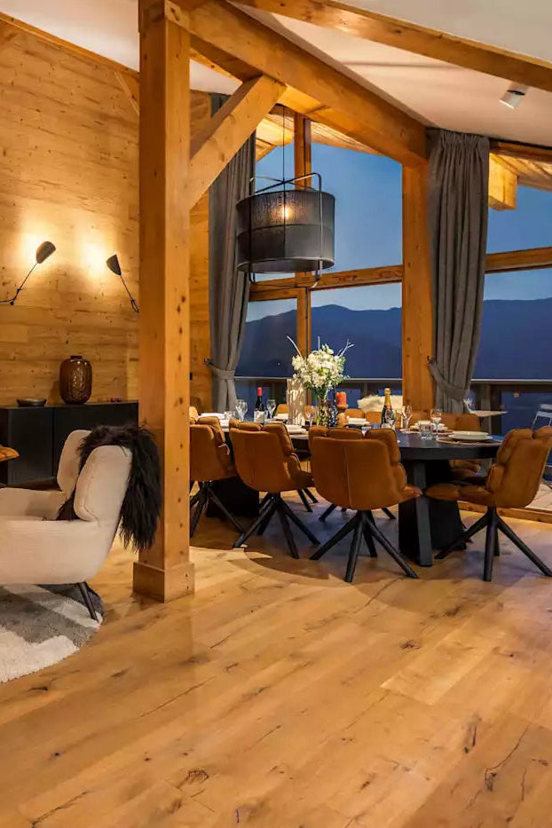 A luxurious mountain chalet interior with warm wood-paneled walls, a dining area with a table and chairs, and a cozy living space with armchairs and a rug, overlooking a mountain view at dusk.