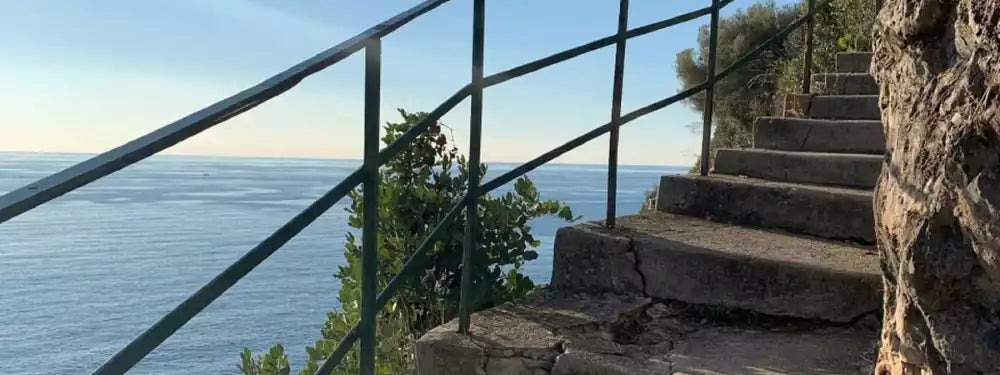 Stone stairs leading up a cliffside overlooking a calm blue sea under a clear sky, with a dark metal railing on the left and a rocky wall on the right.