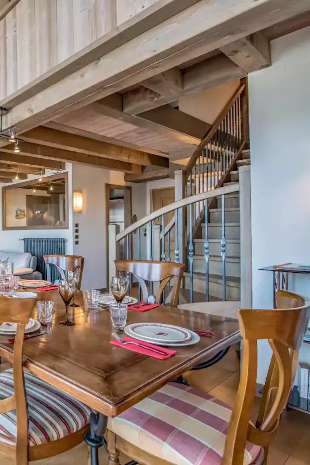 A rustic chalet-style dining room with a wooden dining table and striped upholstered chairs, overlooking a living room with a sofa and a staircase in the background.