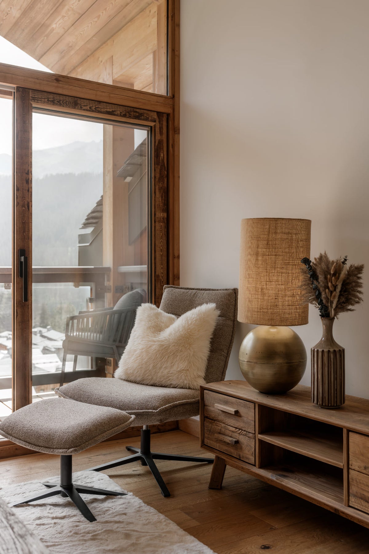A modern lounge chair with a matching ottoman in a warm, rustic room. The chair features a textured taupe-gray fabric and a fluffy cream throw pillow, set next to a wooden sideboard with a woven lampshade table lamp and a vase of dried grass.
