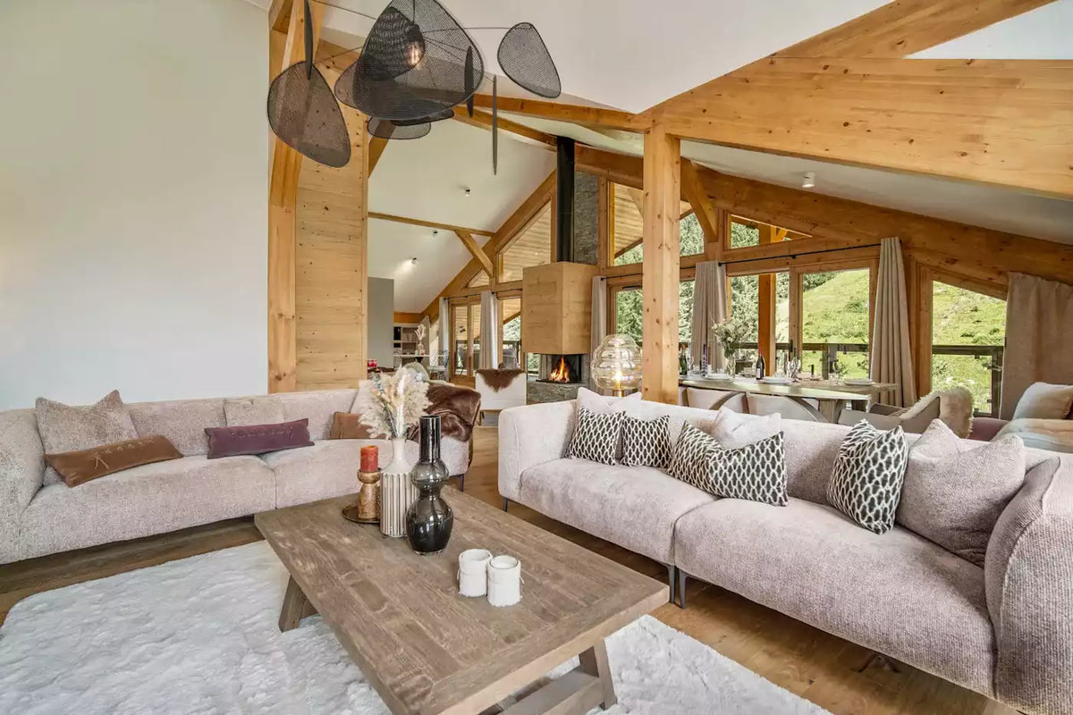 A modern, rustic chalet-style living room featuring two light beige sofas, a wooden coffee table, a large white rug, and a central wood-burning fireplace with exposed wooden beams and tall windows.
