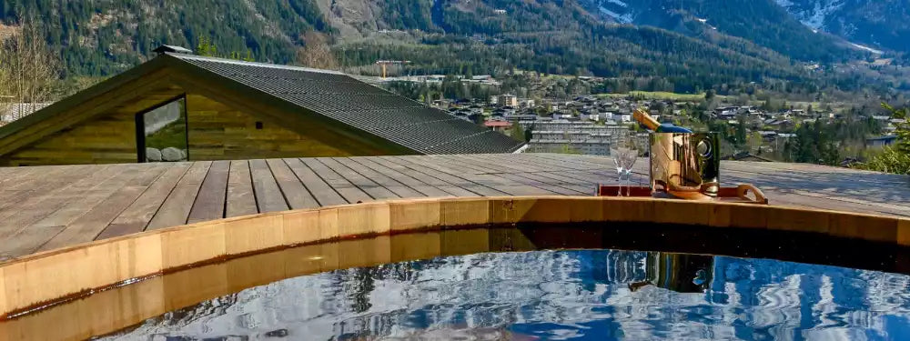 A luxury outdoor terrace with a curved wooden deck, a clear infinity pool reflecting the mountain landscape, and a wine bottle on the deck against a backdrop of pine-covered mountains and a small village.
