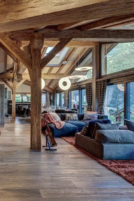 A luxurious mountain chalet living room featuring rustic wooden beams, a large copper-brown fireplace wall with a mounted landscape print, a modern chandelier, and plush blue sofas on patterned rugs.