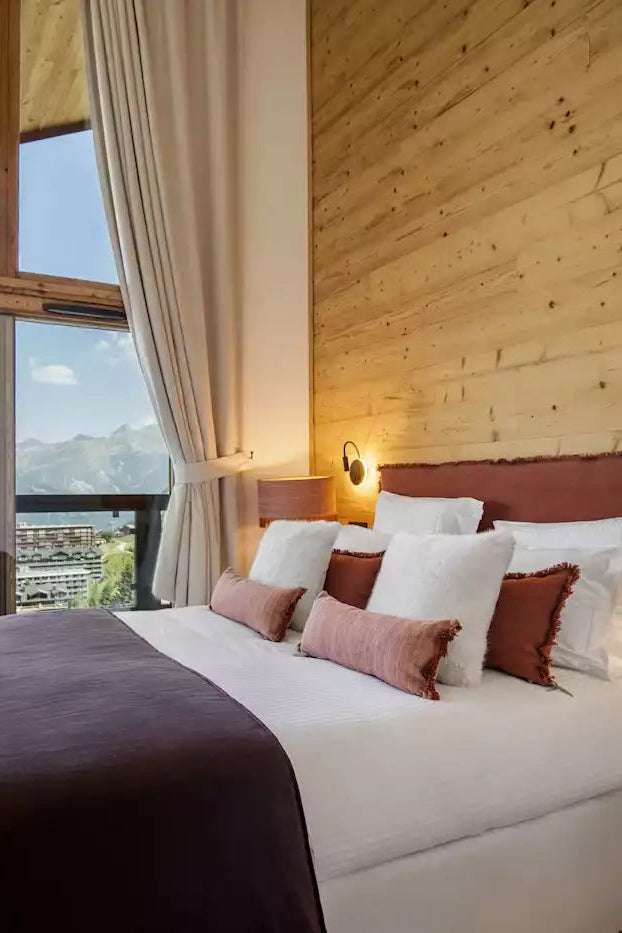 A modern mountain-view bedroom with a large bed dressed in white linens and a dark plum throw, set against a wood-paneled wall. Large windows with beige curtains overlook a scenic lake and mountains, and a wall-mounted TV displays a ski scene.