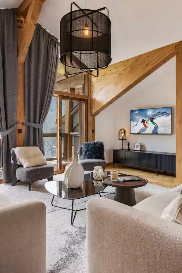 A bright, modern ski chalet living room featuring two cream-colored sofas, a glass coffee table, a wall-mounted TV, and large windows with mountain views.