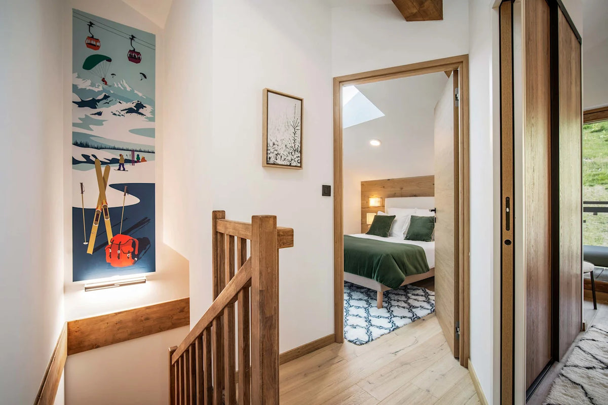 A modern interior hallway with light wood flooring and staircase railings, featuring a large vertical ski-themed wall art print on the left and an open doorway revealing a bedroom with a green bed and patterned rug.