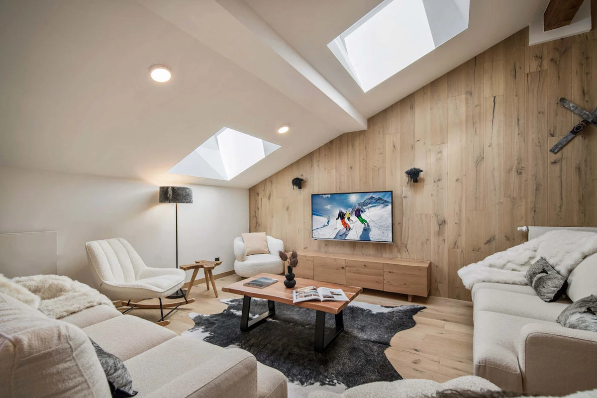 A bright, modern ski chalet living room with light wood paneled walls, multiple skylights, a beige sofa, a white lounge chair, a wooden coffee table, and a TV mounted on the wood wall.