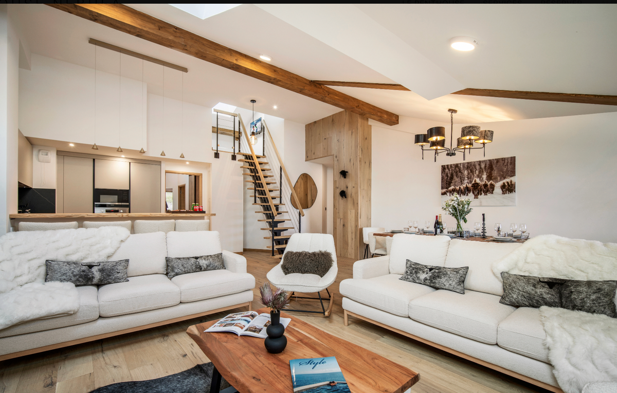 A bright, modern open-plan living room with two large white sofas, a wooden coffee table, a staircase in the background, and exposed wood ceiling beams.