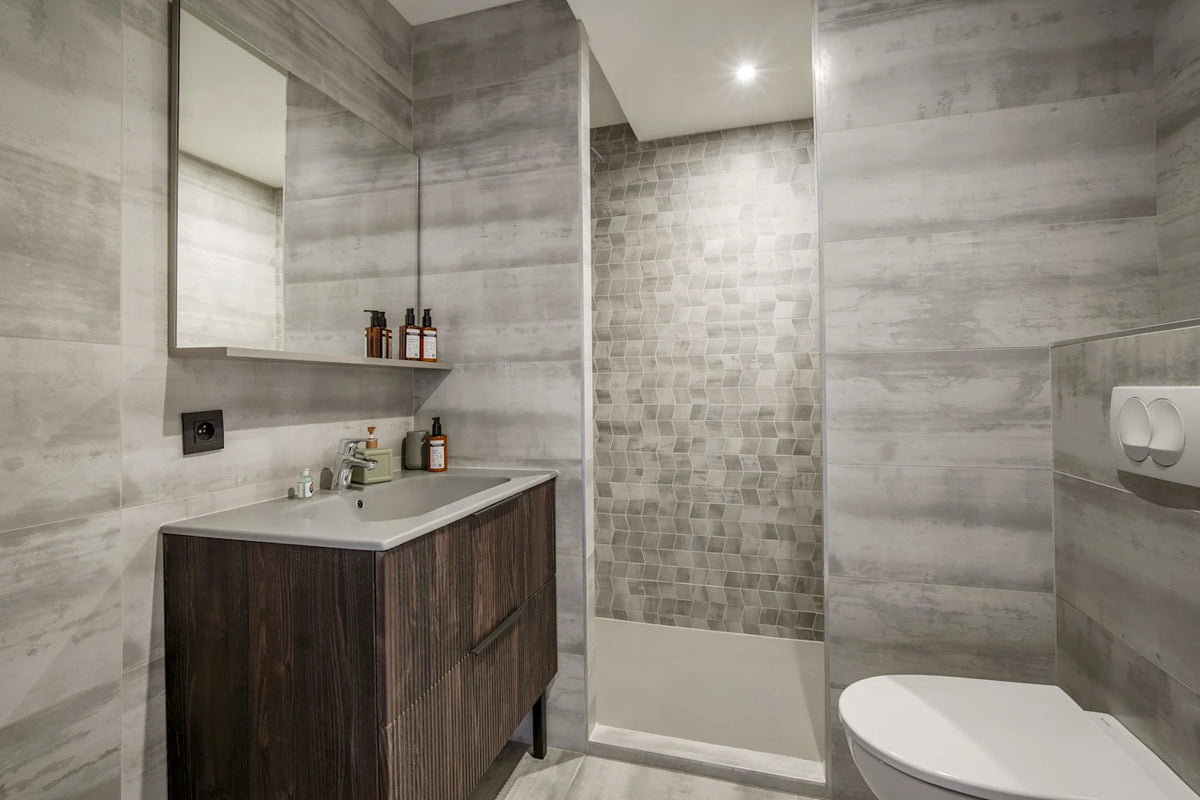 Modern bathroom interior with gray wood-look tiled walls, a dark wood vanity with a white sink, a glass-enclosed shower area with small gray mosaic tiles, and part of a white toilet visible.