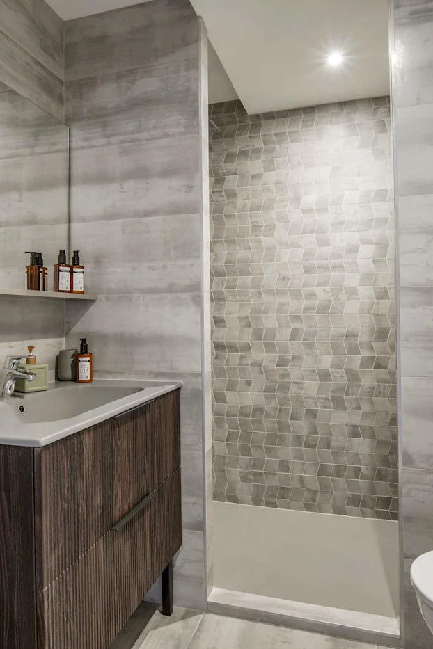 Modern bathroom interior with gray wood-look tiled walls, a dark wood vanity with a white sink, a glass-enclosed shower area with small gray mosaic tiles, and part of a white toilet visible.