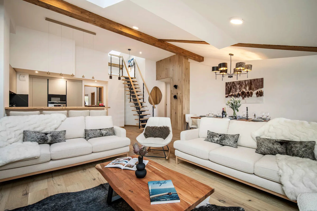 A bright, modern open-plan living room featuring two white sofas with gray patterned pillows, a wooden coffee table, a staircase in the background, and exposed wood ceiling beams.