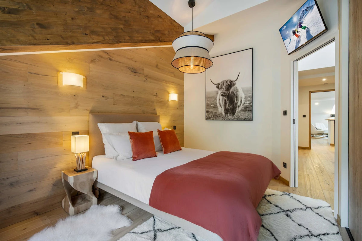 A modern bedroom interior with a neatly made bed, white bedding, rust-orange pillows and throw, a wood-paneled wall, bedside lamps, a hanging pendant light, framed wall art, and an open doorway revealing another room beyond.