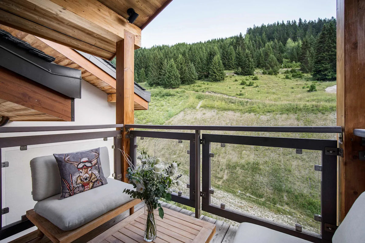 A modern wooden balcony with glass railings, featuring a low wooden bench with gray cushions, a decorative pillow, and a small table with a floral arrangement, overlooking a lush green mountain landscape.