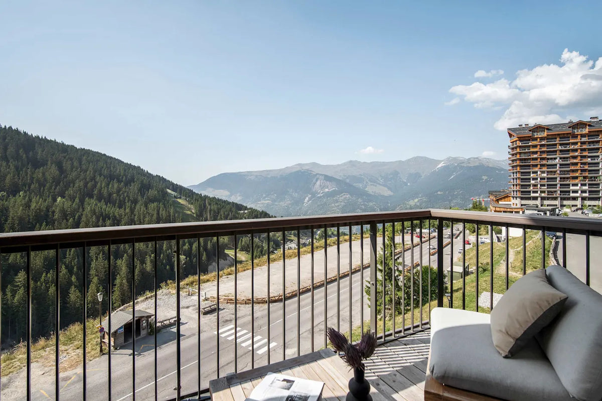 A modern outdoor balcony with a wooden railing and light gray lounge chair, overlooking a scenic mountain road and forested slopes under a blue sky. A tall resort-style building is visible on the right.