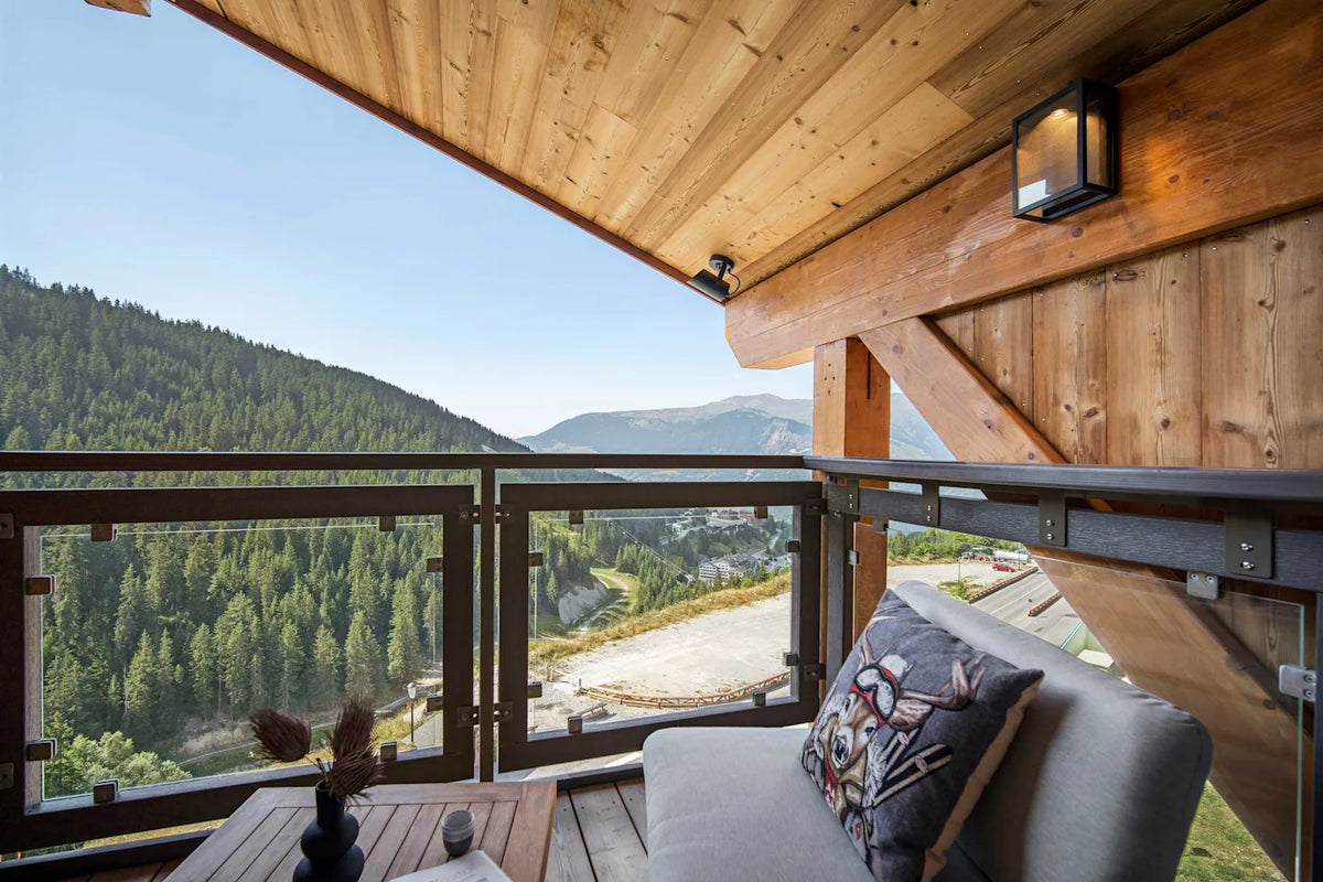A modern wooden chalet balcony with a gray outdoor sofa and decorative pillow, overlooking a lush green mountain valley under a clear blue sky.