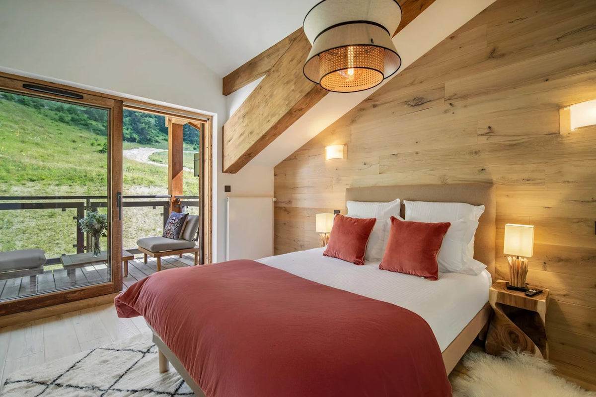 A cozy bedroom interior with a neatly made bed featuring white linens and rust-red pillows and blanket, set against a light wood-paneled wall. Large glass doors open onto a balcony with a grassy outdoor view, and warm wall lights and a hanging pendant lamp illuminate the space.