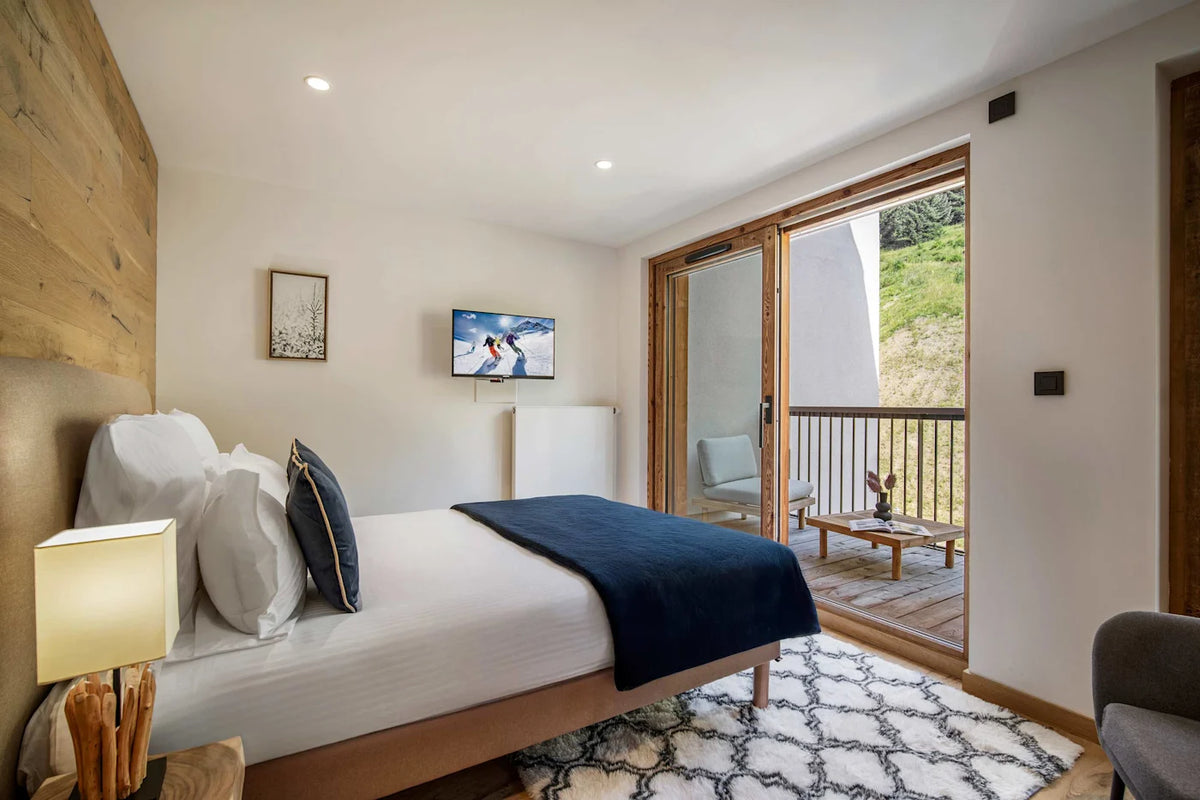 A modern bedroom interior featuring a neatly made bed with white linens and a dark navy throw blanket, set against a light wood headboard. The room has white walls, a wall-mounted TV, a patterned rug, and large sliding glass doors opening onto a wooden balcony with a view of a green outdoor hillside.