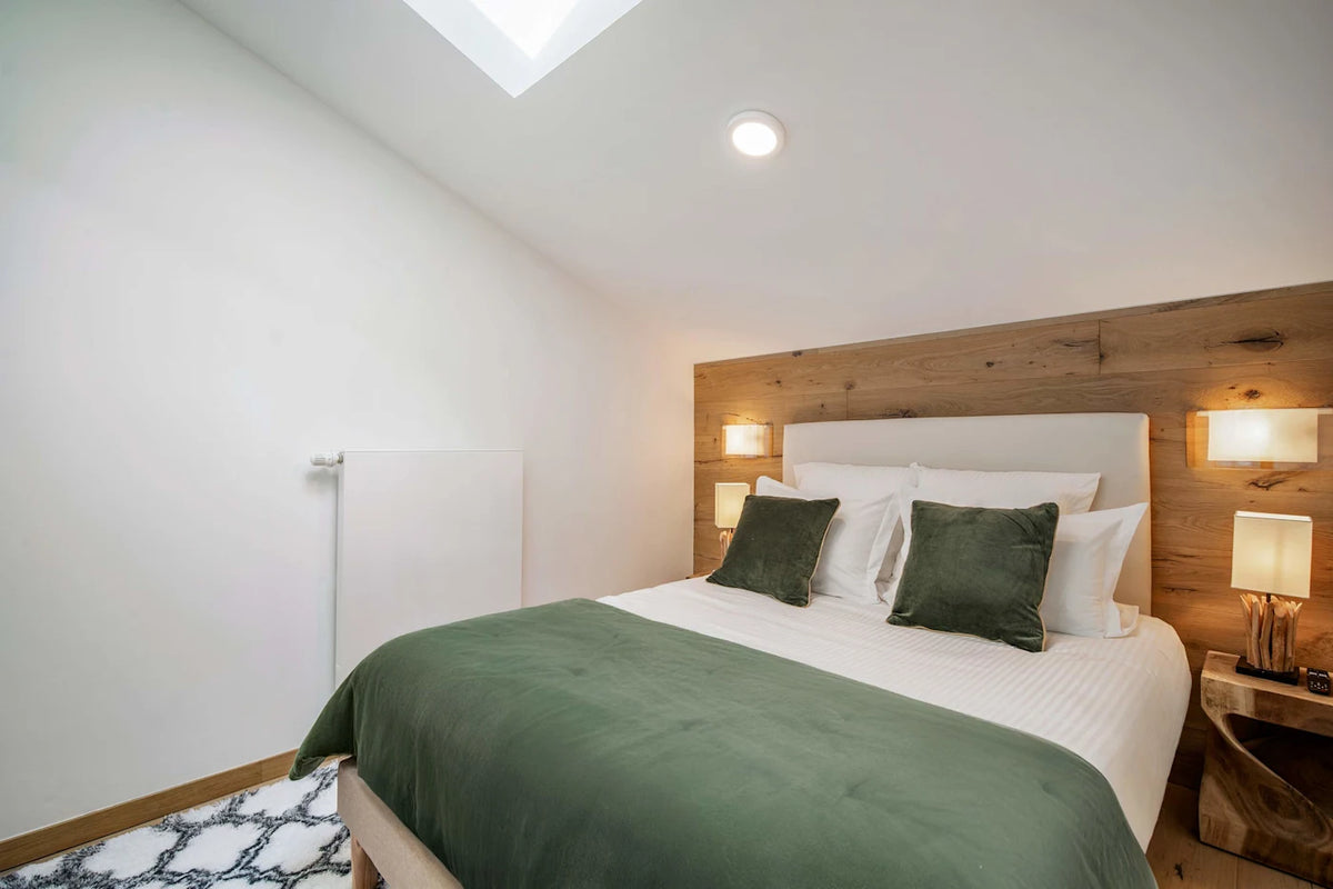 A modern bedroom featuring a neatly made bed with white linens, dark green pillows, and a dark green throw blanket, set against a light wood headboard in a bright room with a skylight.