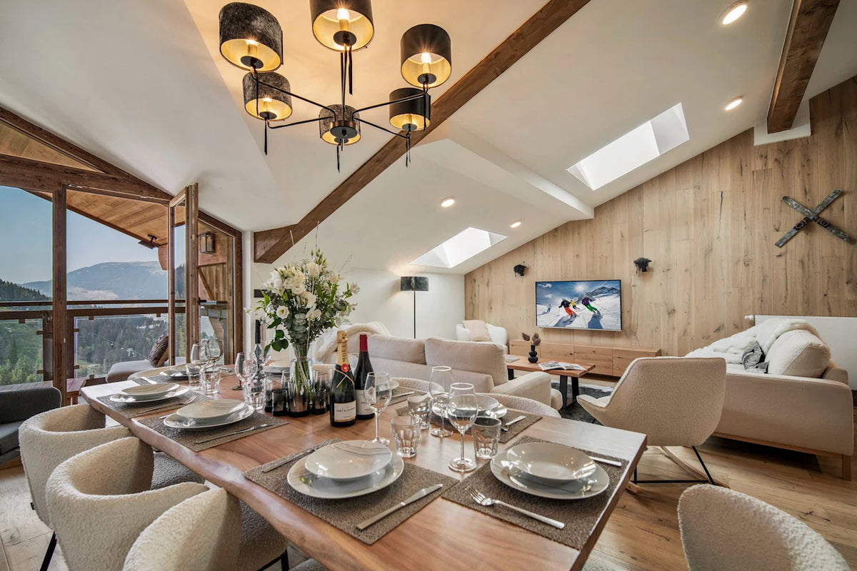 A luxury ski chalet interior featuring a rustic wooden dining table set with white plates and wine bottles, modern cream-colored armchairs, a large multi-light chandelier, a wood-paneled wall with a mounted TV, and a scenic mountain view outside the large windows.