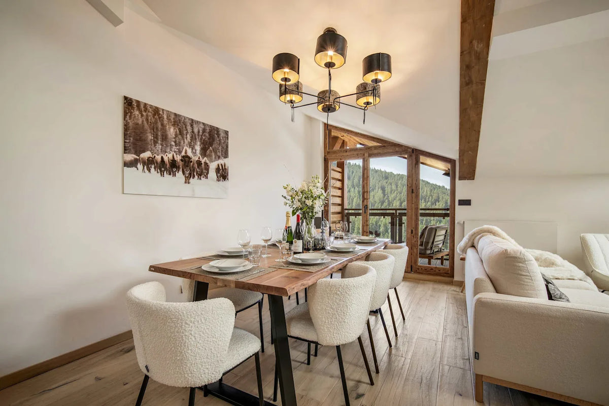 A modern dining room setup in a bright, contemporary chalet-style apartment, featuring a rectangular wooden dining table set with white plates and six textured cream boucle chairs. A multi-light chandelier hangs above, and a large framed landscape print decorates the off-white wall.