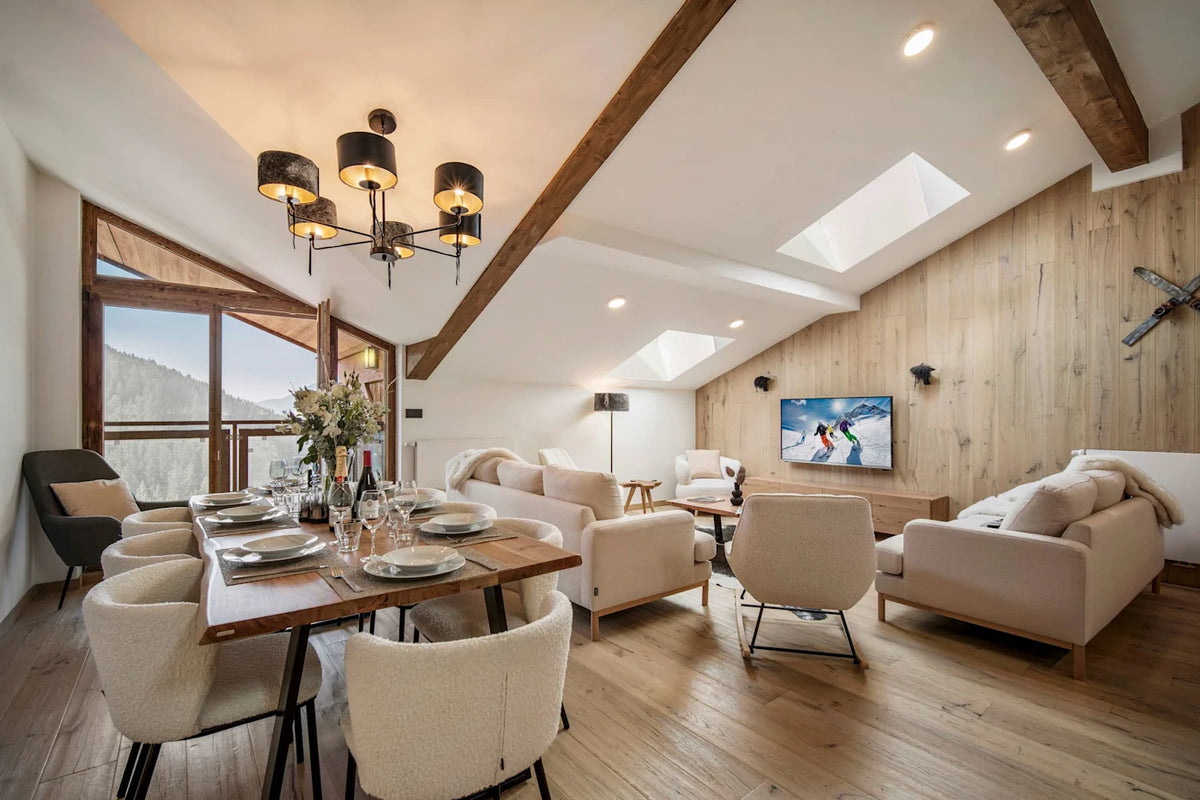 A modern ski resort chalet living and dining area featuring a dining table with cream upholstered chairs, a living room with matching sofas, a wood-paneled wall with a TV, and a slanted ceiling with skylights.