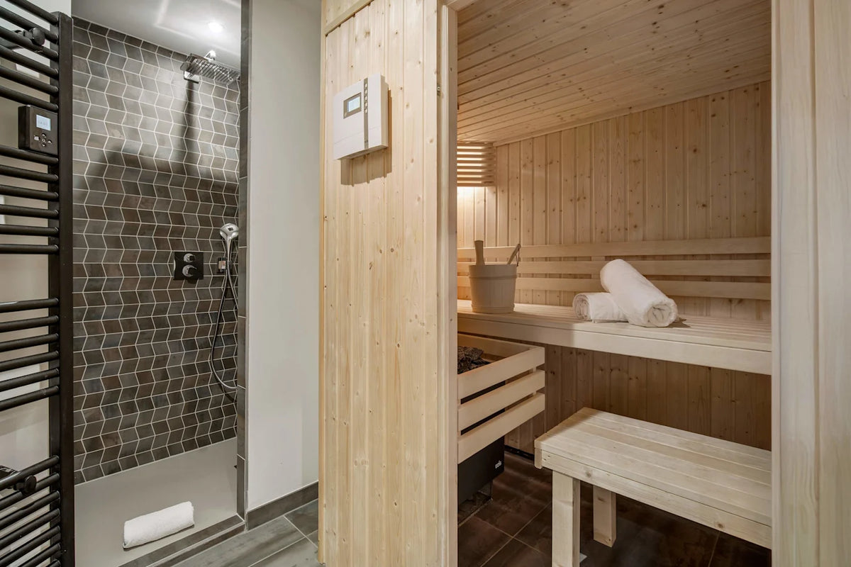 A modern sauna and shower cabin interior with light wood walls and benches, a tiled shower area, and rolled towels on the bench.