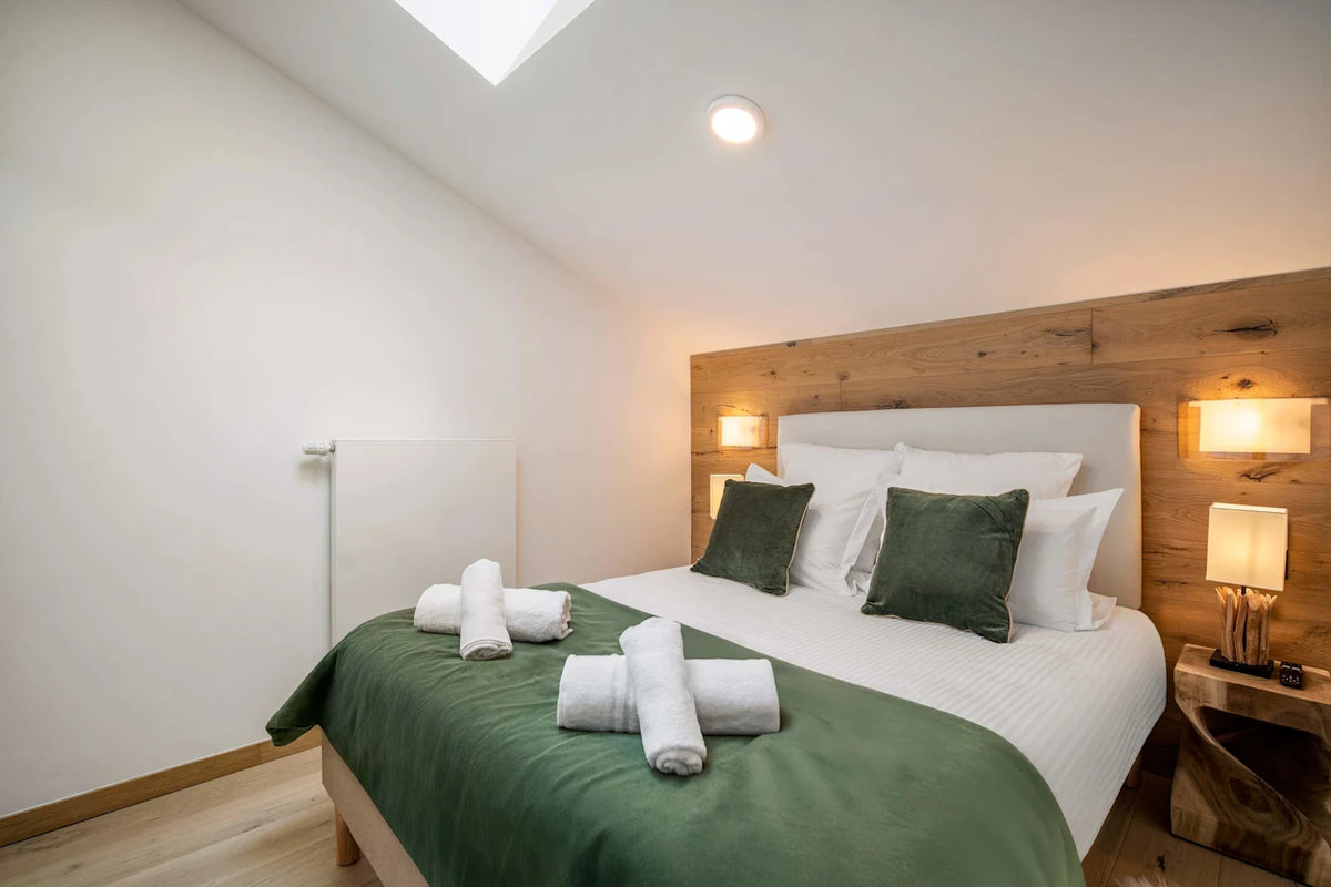A neatly made bed in a modern bedroom with white linens, dark green pillows and throw, a wooden headboard, bedside lamps, and rolled towels on the bedding.
