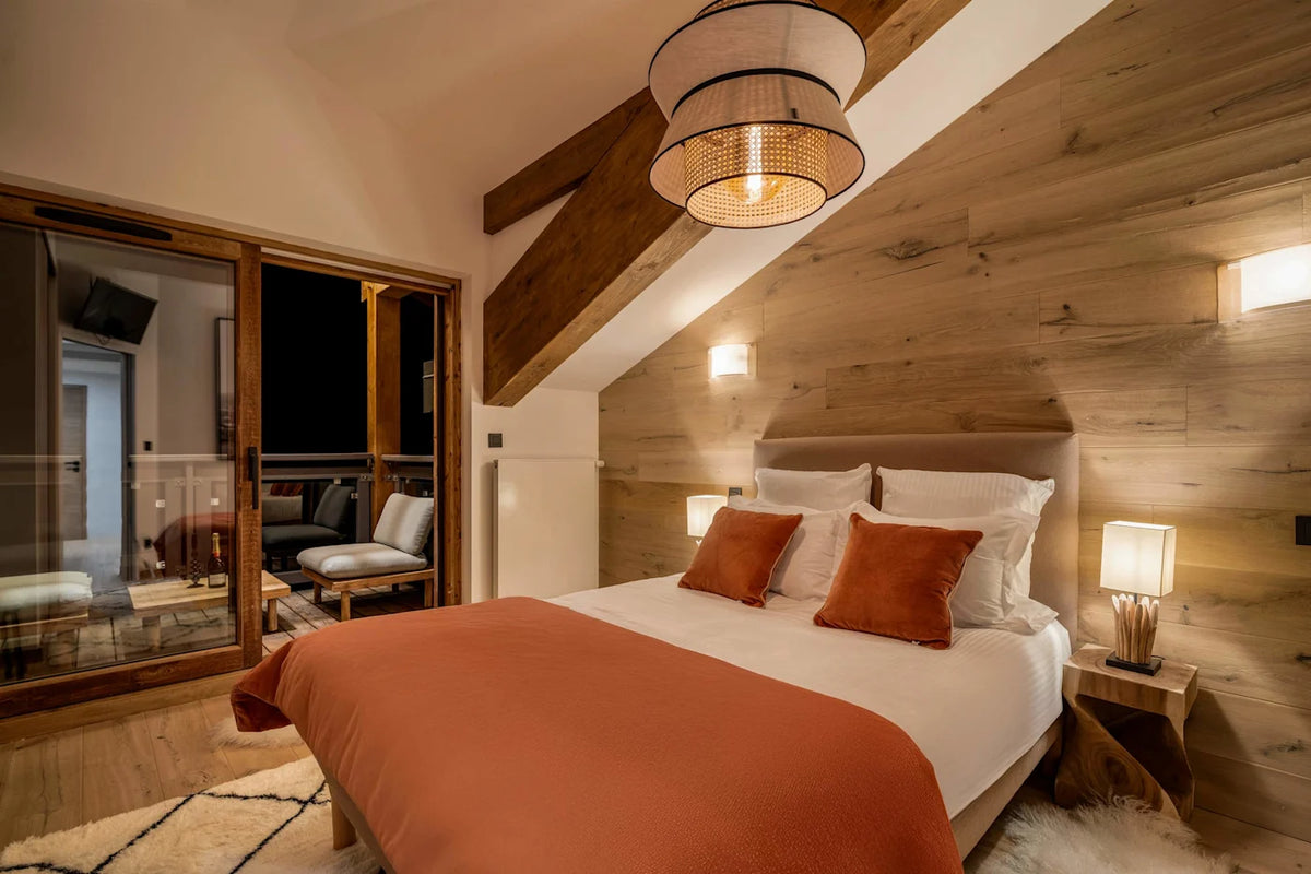 A cozy bedroom interior with a neatly made bed featuring white linens and rust-orange pillows and duvet, set against a light wood-paneled wall with a hanging pendant light and bedside lamps.