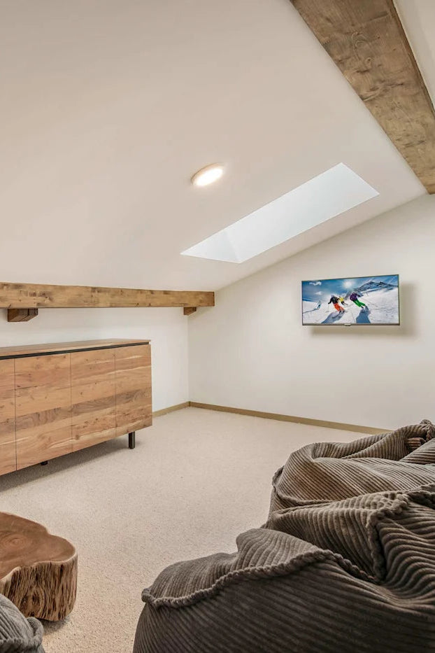 A modern attic room with a wooden credenza, a skylight, exposed wooden beams, and cozy gray corduroy bean bags on a carpeted floor.