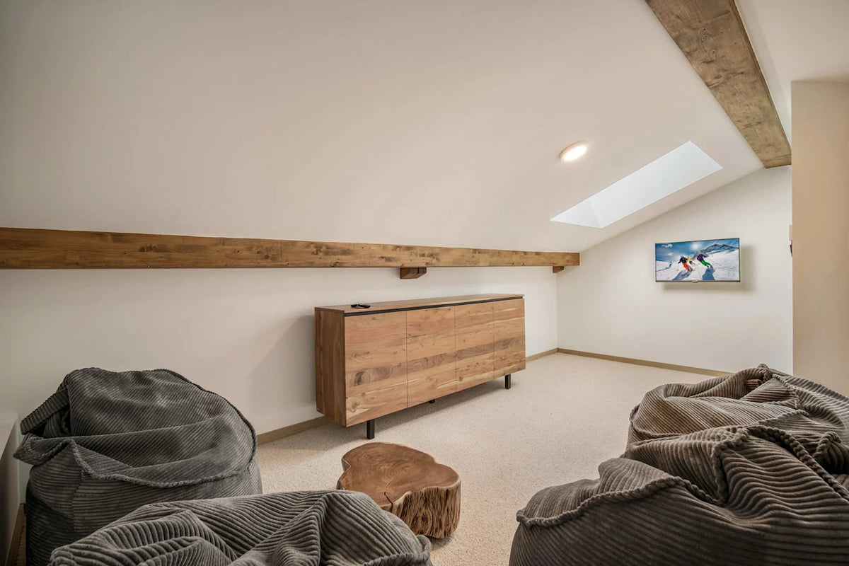 A modern attic room with a wooden credenza, a skylight, exposed wooden beams, and cozy gray corduroy bean bags on a carpeted floor.