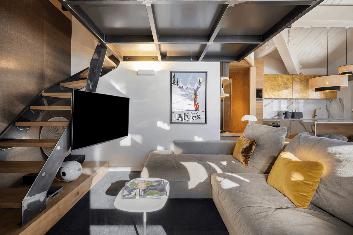 A modern loft-style living room featuring a light gray sectional sofa with yellow accent pillows, a black wall-mounted TV, a wooden staircase with metal railings, and a framed 'Ales' poster on the white wall.