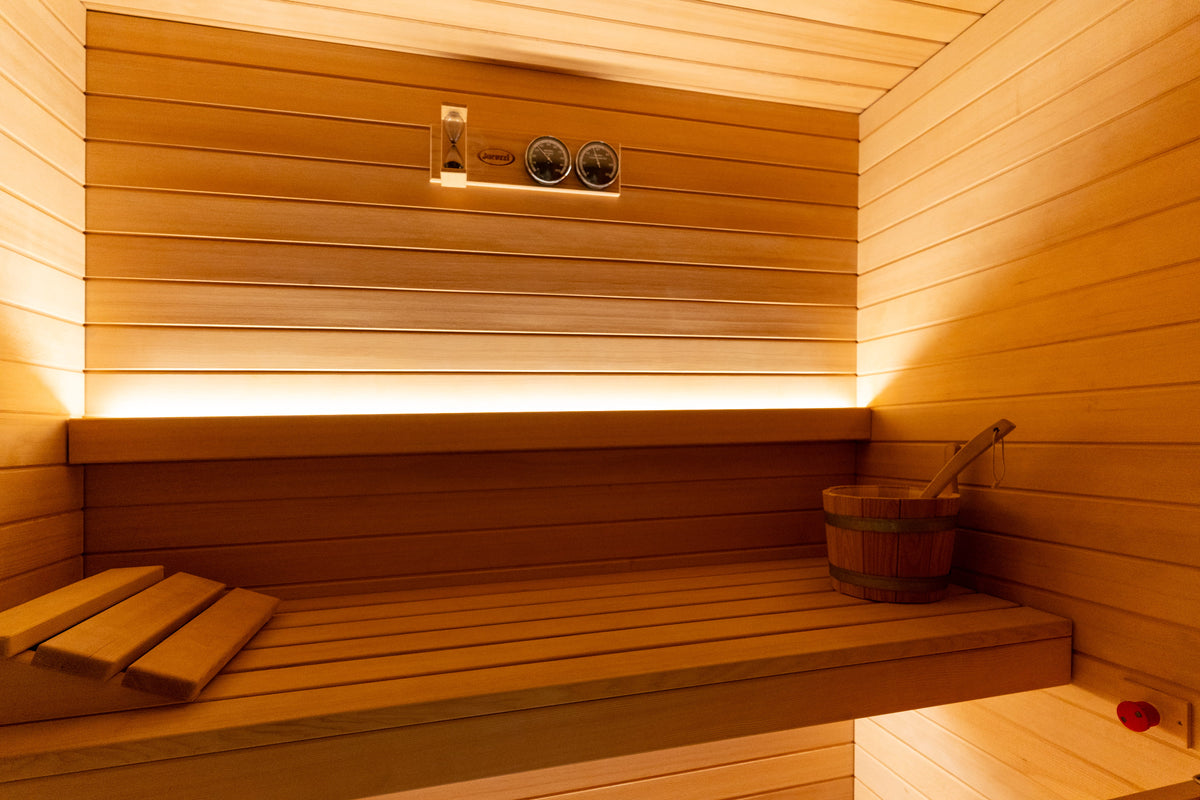 A warm wooden sauna interior with a slatted bench, wall-mounted heating controls, ambient LED lighting, and a wooden bucket with a ladle on the right side.