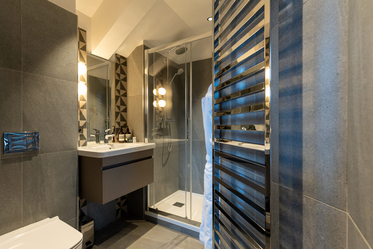 Modern bathroom interior with a glass shower enclosure, floating white sink vanity, dark gray tiled walls, and a tall chrome towel rail.