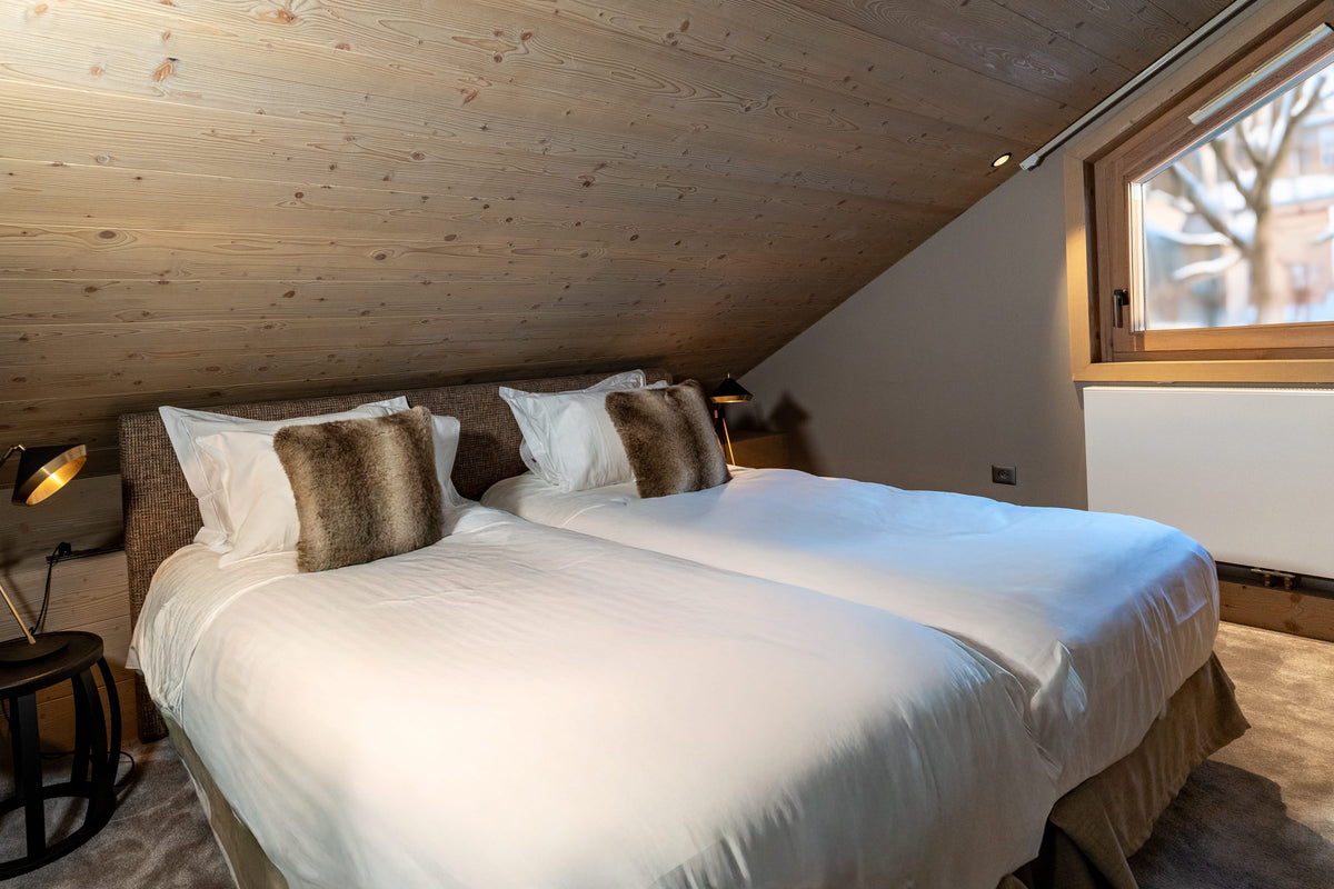A double bed with white bedding and two brown textured accent pillows in a rustic, wood-paneled bedroom with a window.
