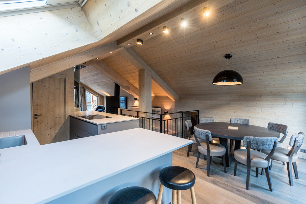 A modern ski chalet interior featuring light wood paneled walls and ceiling, a white kitchen island in the foreground, a dining area with a dark wood table and chairs, and a black pendant light hanging from the ceiling.