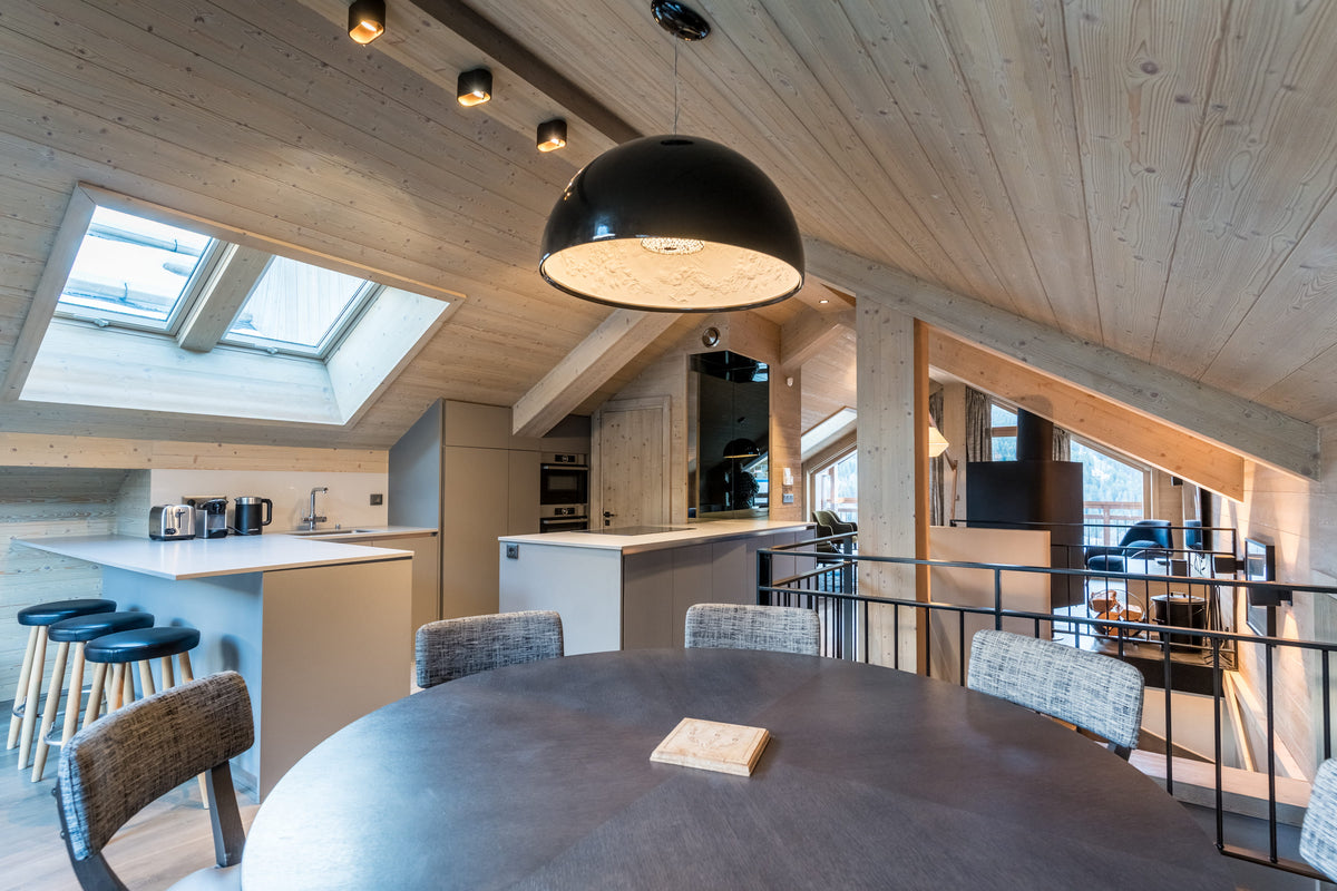 A modern open-plan loft apartment interior with a large round dining table in the foreground, a kitchen island and bar stools in the background, exposed wood ceiling beams, skylights, and a black dome pendant light.