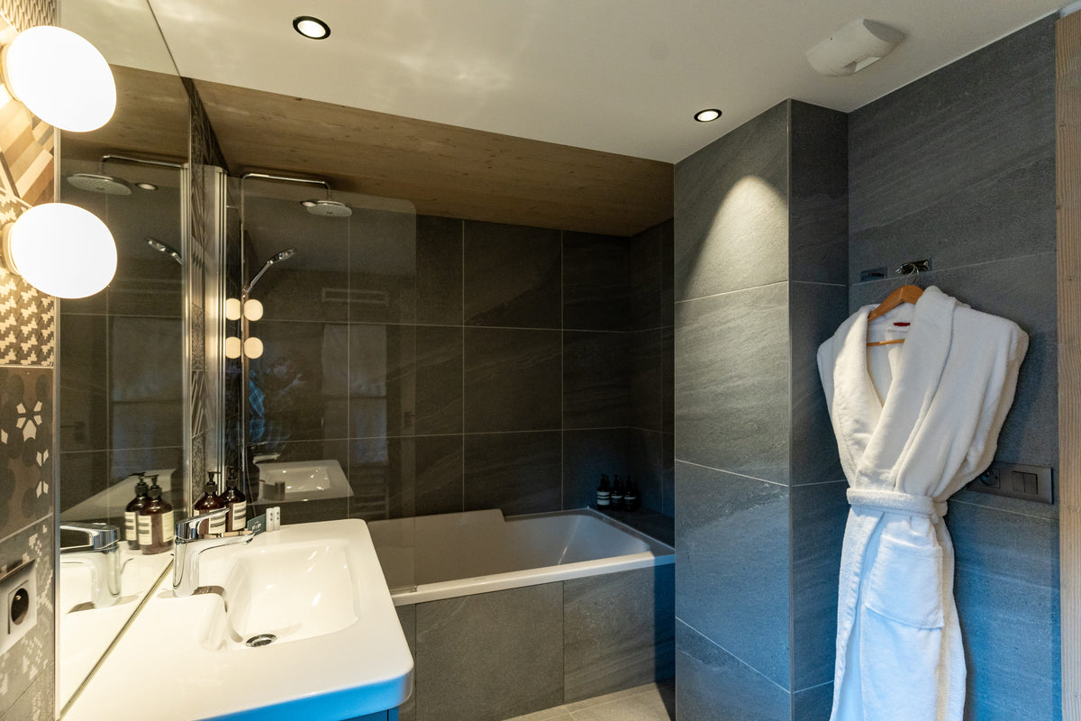 A modern bathroom interior with dark gray tiled walls, a white sink, a bathtub, and a white bathrobe hanging on a hook against the wall.