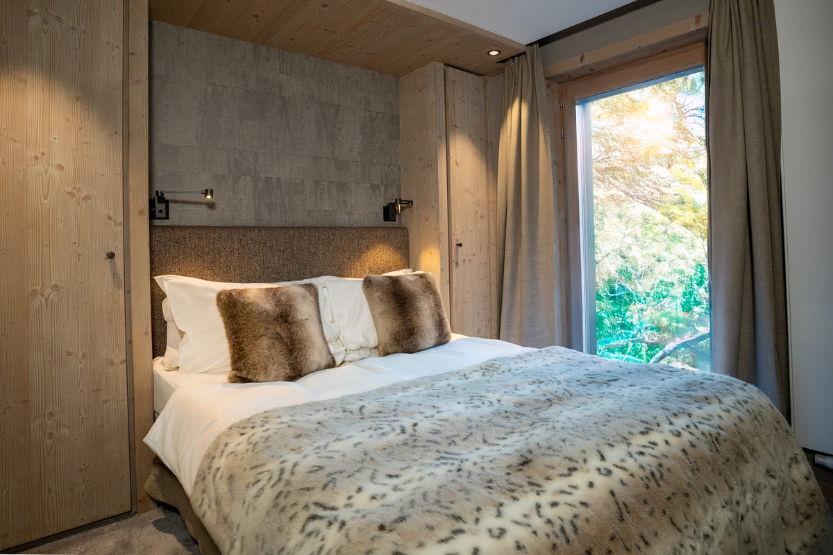 A modern bedroom featuring a large bed dressed in white linens, a beige patterned throw blanket, and two brown faux-fur pillows, set against a wood-paneled wall with a concrete accent wall and a window looking out to green foliage.