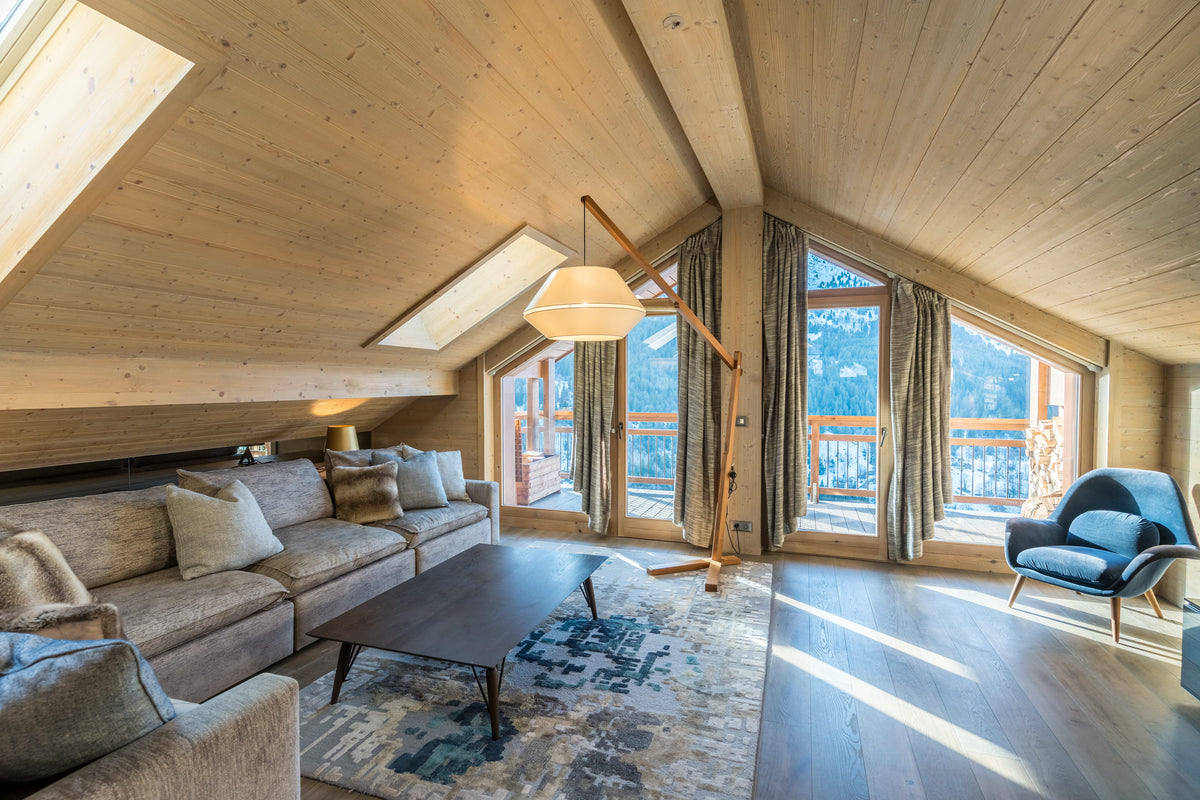 A bright, modern mountain chalet living room with a light wood paneled ceiling and walls, featuring a beige sectional sofa, a dark wood coffee table, a blue accent chair, and large windows with mountain views.