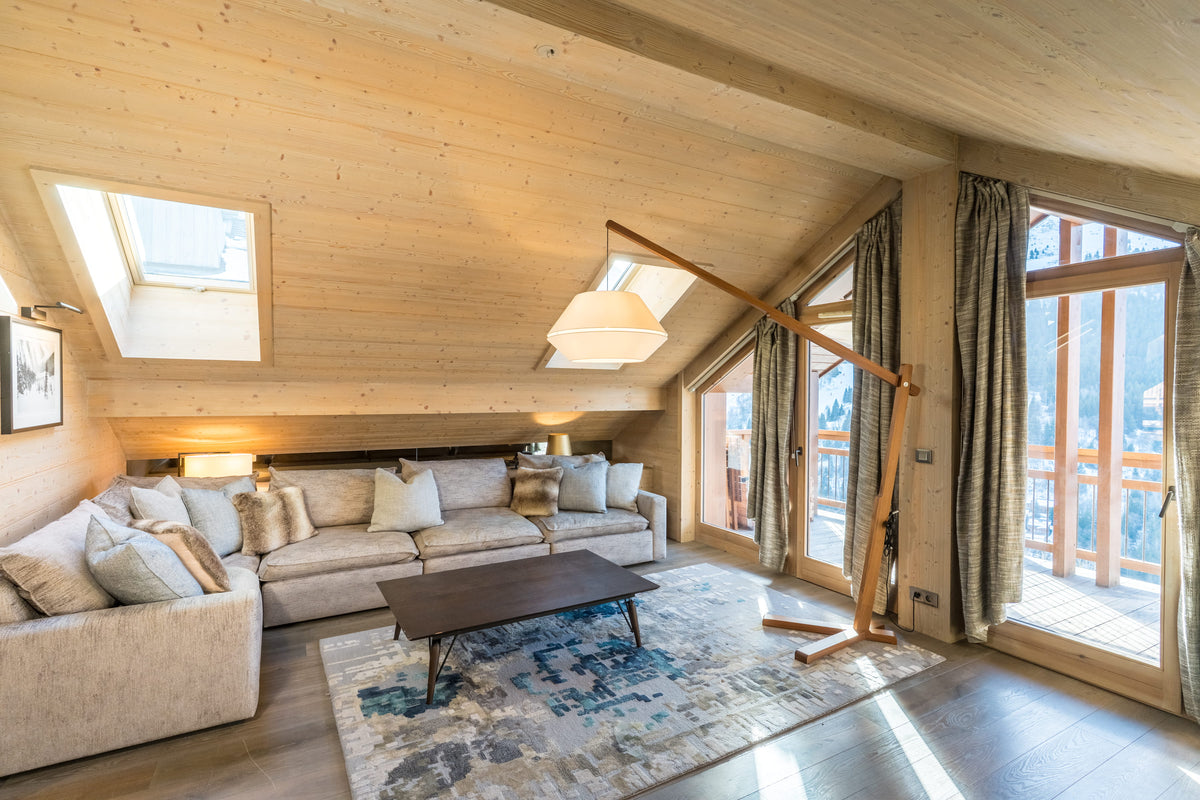 A bright, modern ski chalet living room with a light beige sectional sofa, a dark wood coffee table, a blue and gray patterned area rug, and a large wooden floor lamp against a wood-paneled wall with skylights.