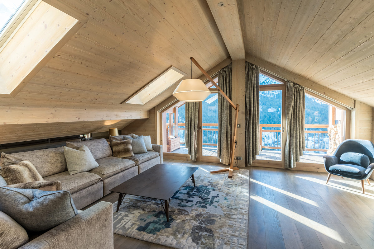 A bright, modern mountain chalet living room with a light wood sloped ceiling, large skylights, a beige sectional sofa, a dark wood coffee table, a patterned rug, and a blue armchair with a scenic view outside the windows.