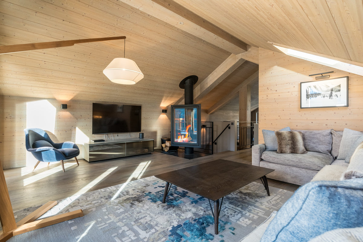 A bright, modern alpine-style living room with light wood walls and ceiling, featuring a gray sofa, blue accent chair, patterned rug, and a black wood-burning fireplace.