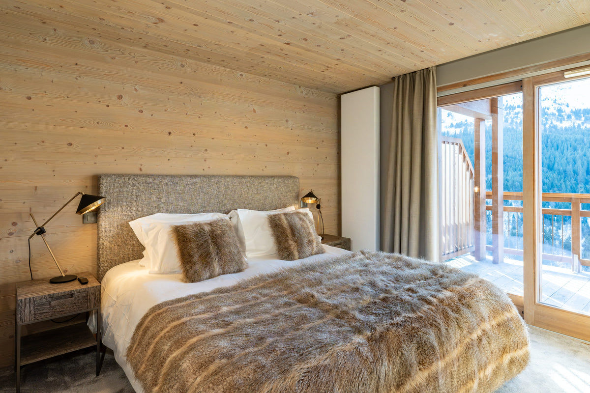 A luxury bedroom featuring a large bed dressed in white linens and a brown faux-fur throw blanket, set against a light wood-paneled wall with a sliding glass door overlooking a mountain landscape.
